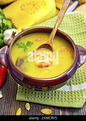 Soupe-purée citrouille avec crevettes et champignons, cuillère dans un bol en argile sur une serviette, persil, gousses de poivron rouge, graines de citrouille, ail sur un bo en bois foncé Banque D'Images