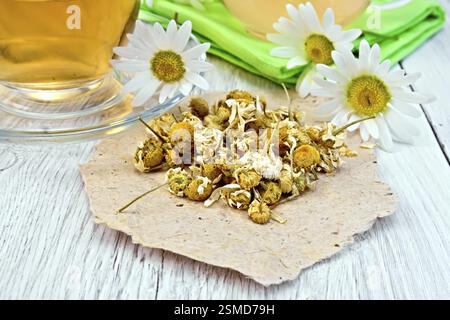 Fleurs de camomille séchées sur papier, plateau en théière en verre sur la serviette et tasse, des fleurs de camomille sur fond de planches en bois Banque D'Images