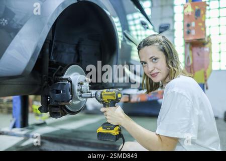 Une femme utilise un outil électrique près du système de freinage d'une voiture, concentrée sur les travaux de réparation dans un atelier Banque D'Images
