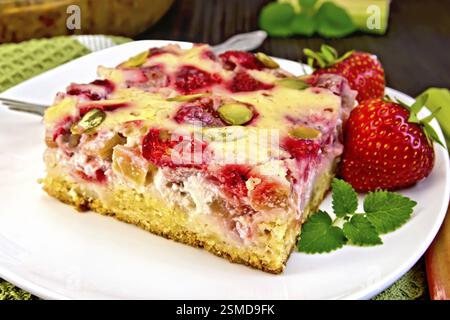 Morceau de tarte avec des fraises, de la rhubarbe et de la sauce à la crème, fourchette, fraise, menthe dans une assiette blanche sur une serviette, tiges de rhubarbe sur un backgroun de planches en bois Banque D'Images