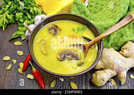 Soupe-purée citrouille avec crevettes et champignons, cuillère dans une tasse brune, serviette, persil, gousses de poivron rouge chaud, graines de citrouille, racine de gingembre sur un bois foncé Banque D'Images