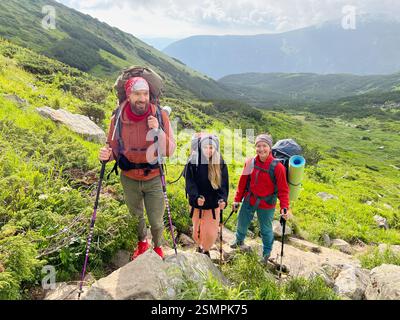 Trois randonneurs, habillés d'équipements de plein air colorés, font une pause sur un sentier rocheux et vert. Le décor pittoresque présente des collines luxuriantes et un ciel partiellement nuageux. Banque D'Images