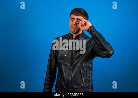 Triste rocker ou motard pleure et essuie les larmes avec la main, debout sur fond bleu Banque D'Images