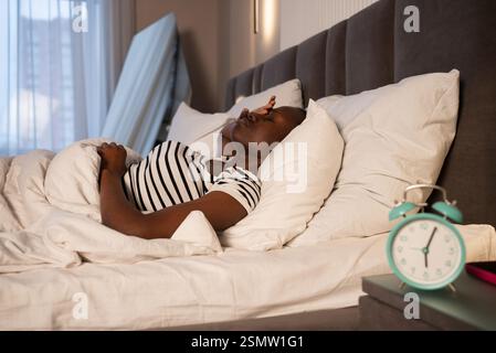 Une femme noire est couchée tôt le matin, couvrant ses yeux avec sa main et souffrant de maux de tête, d'insomnie et d'insomnie Banque D'Images