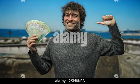 Homme hispanique souriant tout en se pointant vers lui-même et tenant des roubles russes à l'extérieur au bord de la mer par temps clair, célébrant le succès financier avec Banque D'Images