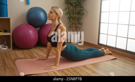 Femme pratiquant le yoga à l'intérieur sur tapis dans le gymnase avec équipement de fitness, montrant calme et concentration, entouré de balles d'exercice et de plantes dans un et lumineux Banque D'Images