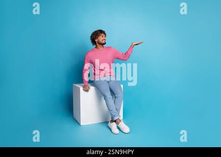 Jeune homme portant un pull rose élégant pose sur un cube sur un fond bleu, montrant un geste amical avec ses mains. Banque D'Images