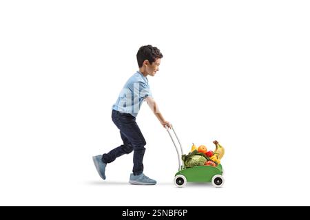 Plan de profil pleine longueur d'un enfant marchant et poussant un chariot à jouets en bois avec des fruits et des légumes isolés sur fond blanc Banque D'Images
