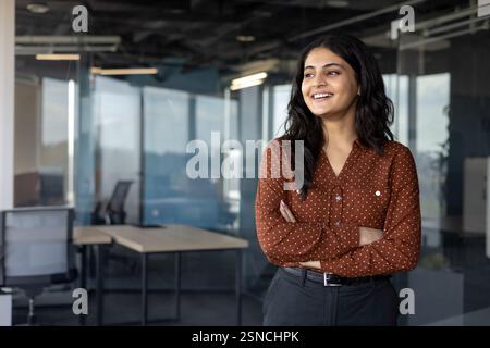 Femme réfléchie et heureuse debout près de la fenêtre à l'intérieur du bureau. Femme d'affaires tenant les bras croisés et regardant sur le côté. Employé de l'entreprise sur le lieu de travail. Banque D'Images