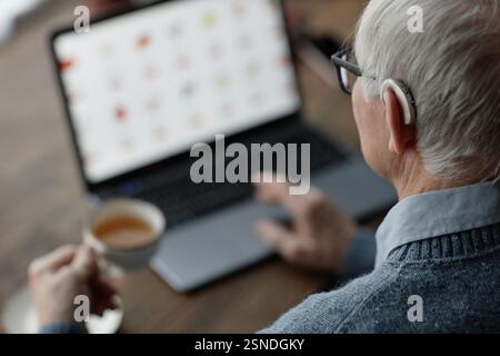 Photo au-dessus de l'épaule d'un homme âgé portant une prothèse auditive derrière l'oreille travaillant sur un ordinateur portable à la maison tout en utilisant une technologie d'assistance, espace de copie Banque D'Images