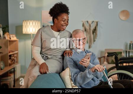 Cliché franc d'une travailleuse sociale noire souriante aidant un homme âgé à utiliser la technologie tout en regardant une vidéo au téléphone ensemble dans le salon Banque D'Images