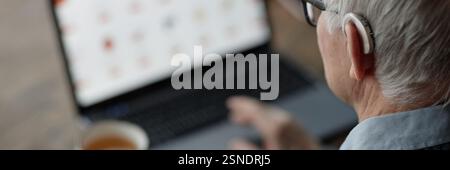 Image d'en-tête de site Web d'un homme âgé portant une aide auditive derrière l'oreille en utilisant un ordinateur portable à la maison avec l'aide de la technologie d'assistance, espace de copie Banque D'Images