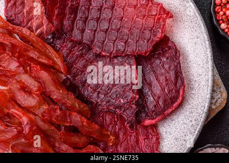 Tranches de bœuf séchées, saccadées, avec saveur de cerise ou de prune, sel et épices sur un fond de béton foncé Banque D'Images