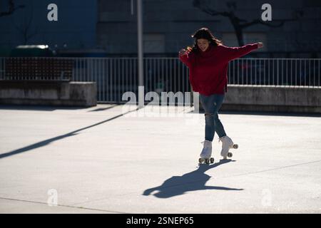 Jeune femme avec un Jean déchiré et un pull rouge glisse gracieusement sur des patins à roulettes dans un parc de la ville, embrassant la joie de l'activité de plein air et de l'exercice Banque D'Images