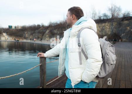 Bel homme avec sac à dos sur une promenade en bois au bord d'un lac, entouré de falaises rocheuses et d'arbres d'automne. Homme touristique en plein air en hiver Banque D'Images