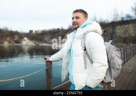 Bel homme avec sac à dos sur une promenade en bois au bord d'un lac, entouré de falaises rocheuses et d'arbres d'automne. Homme touristique en plein air en hiver Banque D'Images