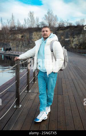 Bel homme avec sac à dos sur une promenade en bois au bord d'un lac, entouré de falaises rocheuses et d'arbres d'automne. Homme touristique en plein air en hiver Banque D'Images