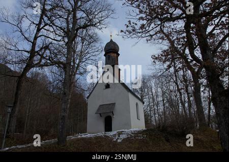 Une petite église blanche avec un clocher se dresse au milieu d'une forêt en Allemagne, Munich. De grands arbres entourent l'église et la neige recouvre son toit. T Banque D'Images