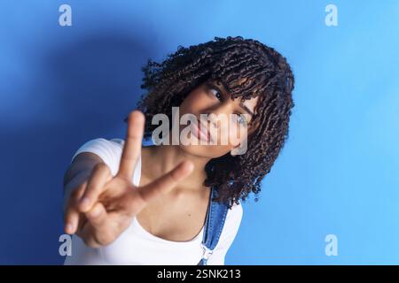 Jeune femme aux cheveux bouclés faisant un signe de victoire avec ses doigts sur un fond bleu vif, rayonnant confiance et positivité Banque D'Images