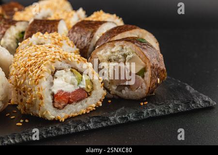 Les rouleaux de sushi colorés sont agencés avec art sur une assiette en pierre sombre, présentant une variété de garnitures comme le poisson et le fromage à la crème, surmonté de graines de sésame Banque D'Images