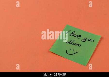Une note collante verte est placée sur une surface orange vif, exprimant l'amour et la gratitude avec les mots je t'aime maman et un visage souriant dessiné ci-dessous. Banque D'Images