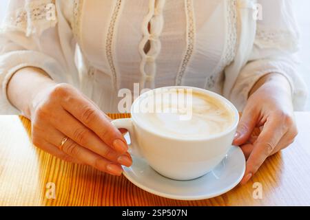 Femme dans une robe de dentelle blanche tient une tasse de cappuccino derrière un café en bois. Banque D'Images