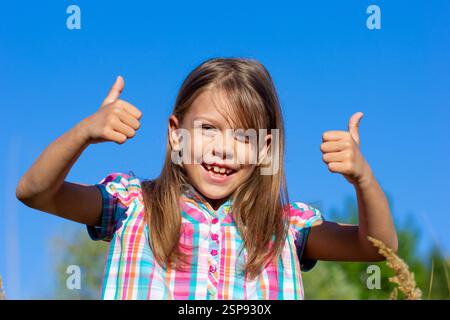 Portrait d'un enfant caucasien souriant 7 8 ans montrant les pouces vers le haut sur le ciel bleu Banque D'Images