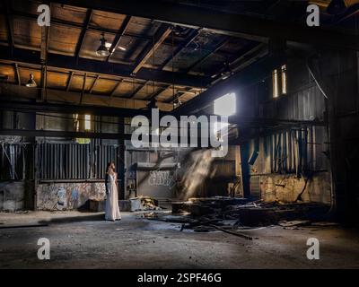 Femme debout dans la lumière du soleil brisant à travers la fenêtre cassée dans l'usine abandonnée sale, Conshohocken, Pennsylvanie, États-Unis Banque D'Images