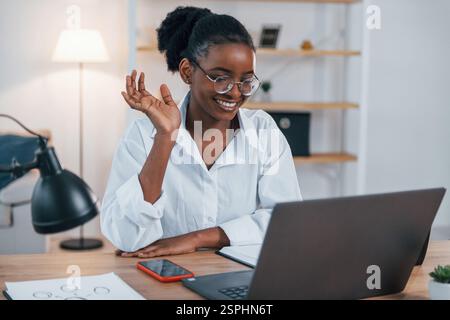Avoir une réunion en ligne. La jeune femme afro-américaine en chemise blanche est à la maison. Banque D'Images