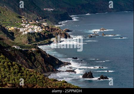 Une vue panoramique sur la côte avec des falaises accidentées, des vagues qui s'écrasent contre des rochers et un petit village perché sur la colline. Le paysage est luxuriant Banque D'Images