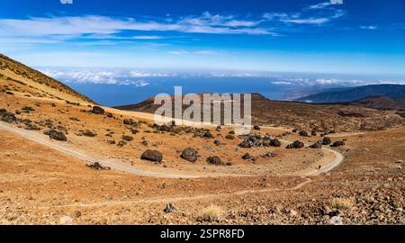 Une vue panoramique d'un chemin de terre sinueux à travers un paysage aride avec un terrain rocheux, menant à une vue lointaine sur l'océan sous un ciel bleu clair avec s. Banque D'Images