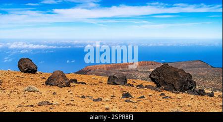 Une vue panoramique sur un paysage rocheux avec de grands rochers dispersés sur un terrain sec, surplombant un vaste océan sous un ciel bleu vif avec des nuages tortueux Banque D'Images