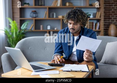 Homme travaillant à la maison avec des documents, des factures et des chèques. Utilisation d'un ordinateur portable et d'une calculatrice pour calculer les factures de services publics, loyer. Assis sur le canapé dans le salon de l'appartement. Banque D'Images