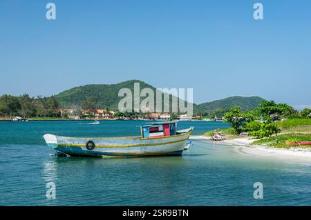 Un petit bateau de pêche ancré sur la rive nord-est du canal Itajuru avec une végétation verte et des maisons lointaines sous un ciel bleu ensoleillé après-midi d'été. Banque D'Images
