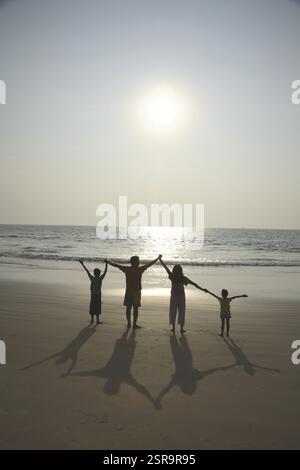 Parent indien sud-asiatique et enfants tenant les mains face au soleil avec de longues ombres derrière eux sur le sable à Seashore, Shiroda, Dist. Sindhudurga, Maharas Banque D'Images