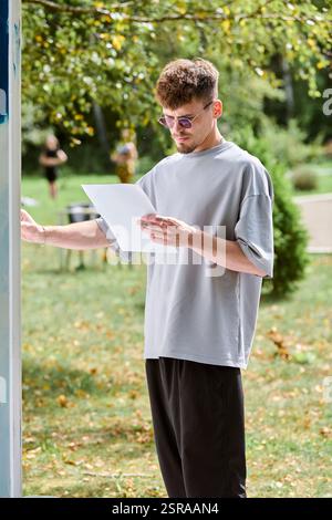 Jeune artiste masculin adulte concentré avec des lunettes dessinant sur une grande toile blanche à l'extérieur, entouré d'une végétation luxuriante. Filtre la lumière du soleil douce Banque D'Images
