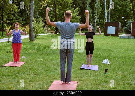Jeune homme adulte dirigeant session de yoga pour femme blanche mature et jeune femme blanche adulte. Parc vert environnant, avec éclairage doux et atmosphère décontractée Banque D'Images