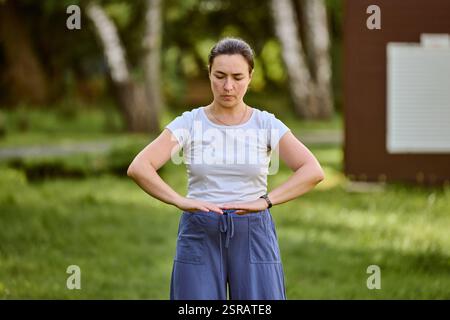 Jeune femme adulte avec les cheveux foncés, pratiquant le qigong dans le parc ensoleillé. Expression focalisée, bras formant cercle. Fond d'arbres et d'herbe verte douce Banque D'Images