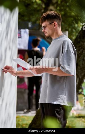 Jeune artiste masculin adulte concentré avec des lunettes dessinant sur une grande toile blanche à l'extérieur, entouré d'une végétation luxuriante. Filtre la lumière du soleil douce Banque D'Images