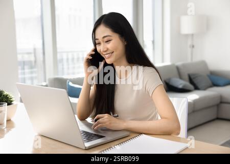 Femme asiatique assise au bureau, souriante, parler principal sur smartphone Banque D'Images