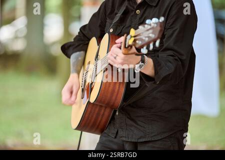 Kaliningrad, Russie - 10.08.2024 - jeune musicien masculin strums guitare acoustique dans un parc ensoleillé. Portant une chemise noire, concentrez-vous sur les mains et la guitare. Natu doux Banque D'Images