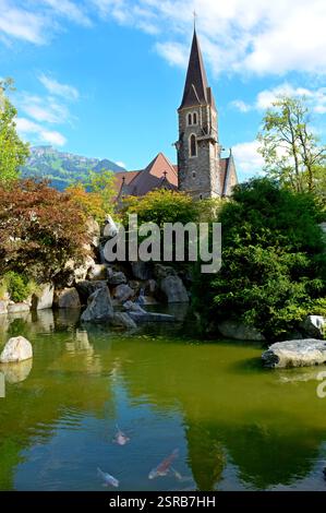 Une église historique avec un clocher pointu surplombe un étang tranquille à carpes koï avec une végétation luxuriante, des cascades et des reflets dans l'eau. Banque D'Images