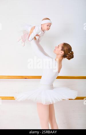 Jeune adulte ballerine féminine avec cheveux blonds soulève ludique petit bébé femelle dans le studio de ballet. Un éclairage lumineux met en valeur leurs expressions joyeuses Banque D'Images