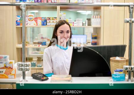 Jeune pharmacienne adulte avec de longs cheveux bruns se tient souriant derrière le comptoir dans une pharmacie bien éclairée, entourée d'étagères de médicaments. Professio Banque D'Images