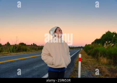 Heureuse jeune femme asiatique vêtue d'un sourire de manteau confortable sur le bord de la route à l'aube à la Nouvelle-Zélande Banque D'Images