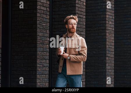 Pause café. Homme élégant avec la barbe dans la veste de couleur kaki et en jeans est à l'extérieur près du bâtiment. Banque D'Images