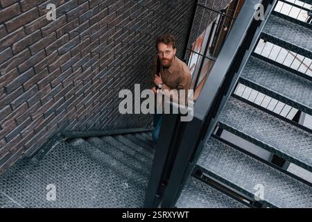 Recherche en haut. Homme en veste de couleur kaki et en jeans est sur les escaliers sur le bâtiment à l'extérieur. Banque D'Images