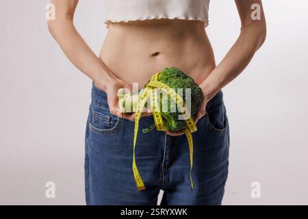 Femme mince tenant le brocoli enveloppé dans un ruban à mesurer, symbolisant une alimentation saine, la perte de poids, l'alimentation et la nutrition sur fond blanc. Banque D'Images