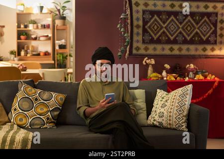 Homme portant une tenue traditionnelle assis sur le canapé dans le salon confortable tout en lisant des messages sur le téléphone. Le fond comprend des étagères, des plantes et des tapisseries accrochées au mur Banque D'Images