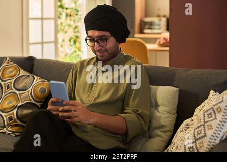 Homme souriant portant turban assis sur un canapé tenant un smartphone, entouré de coussins et de décoration, créant une atmosphère chaleureuse Banque D'Images